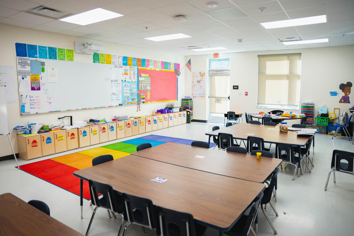 Bright elementary school classroom with colorful alphabet cards above a whiteboard, low cabinets, and tables with black chairs around them.