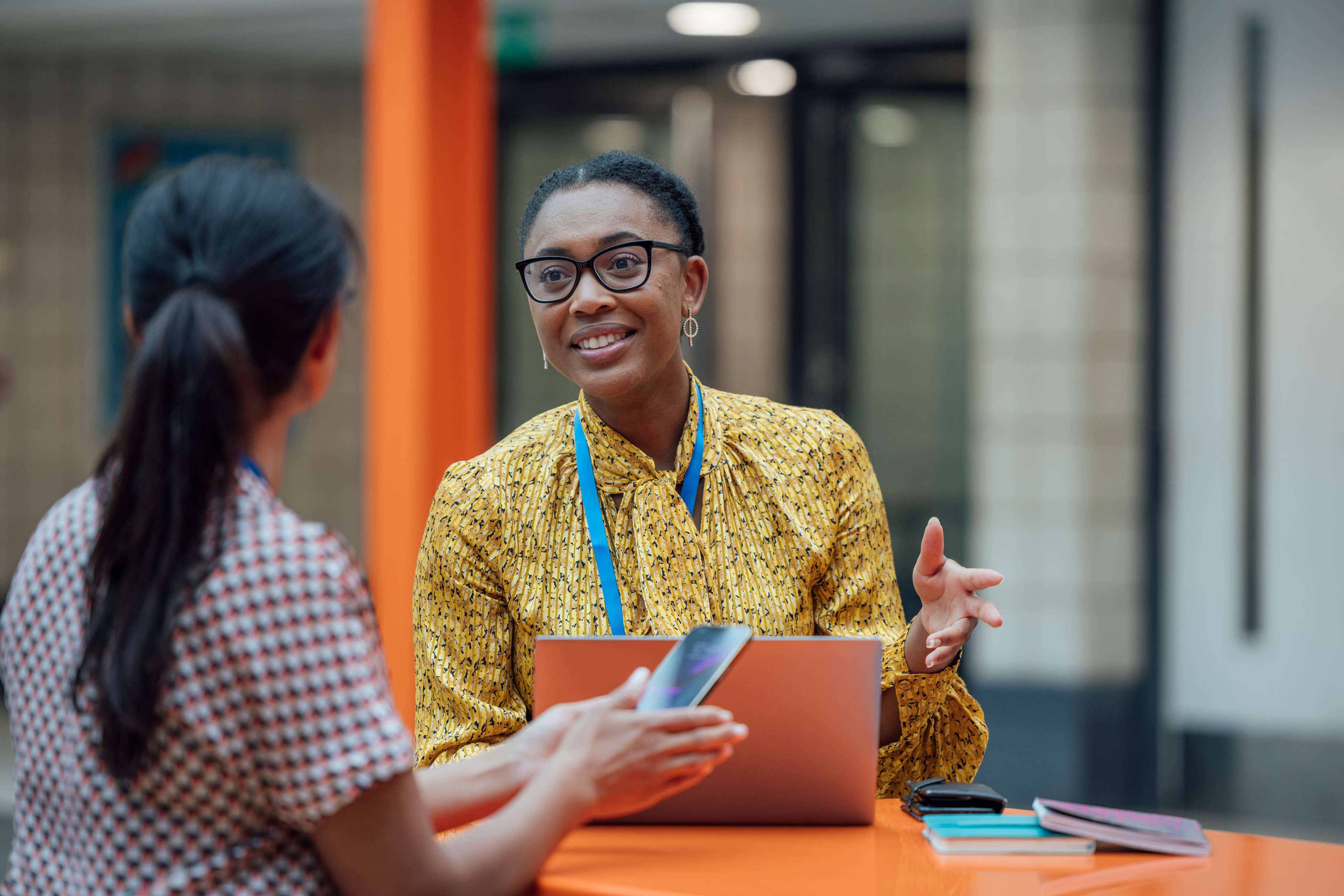 Two women having a discussion at an orange table, one holding a smartphone and the other gesturing near a closed laptop.