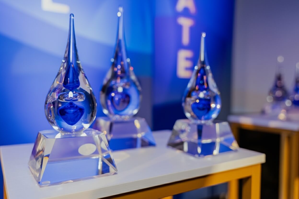 Three glass teardrop-shaped awards with blue heart designs inside, displayed on a white table with a blue background.