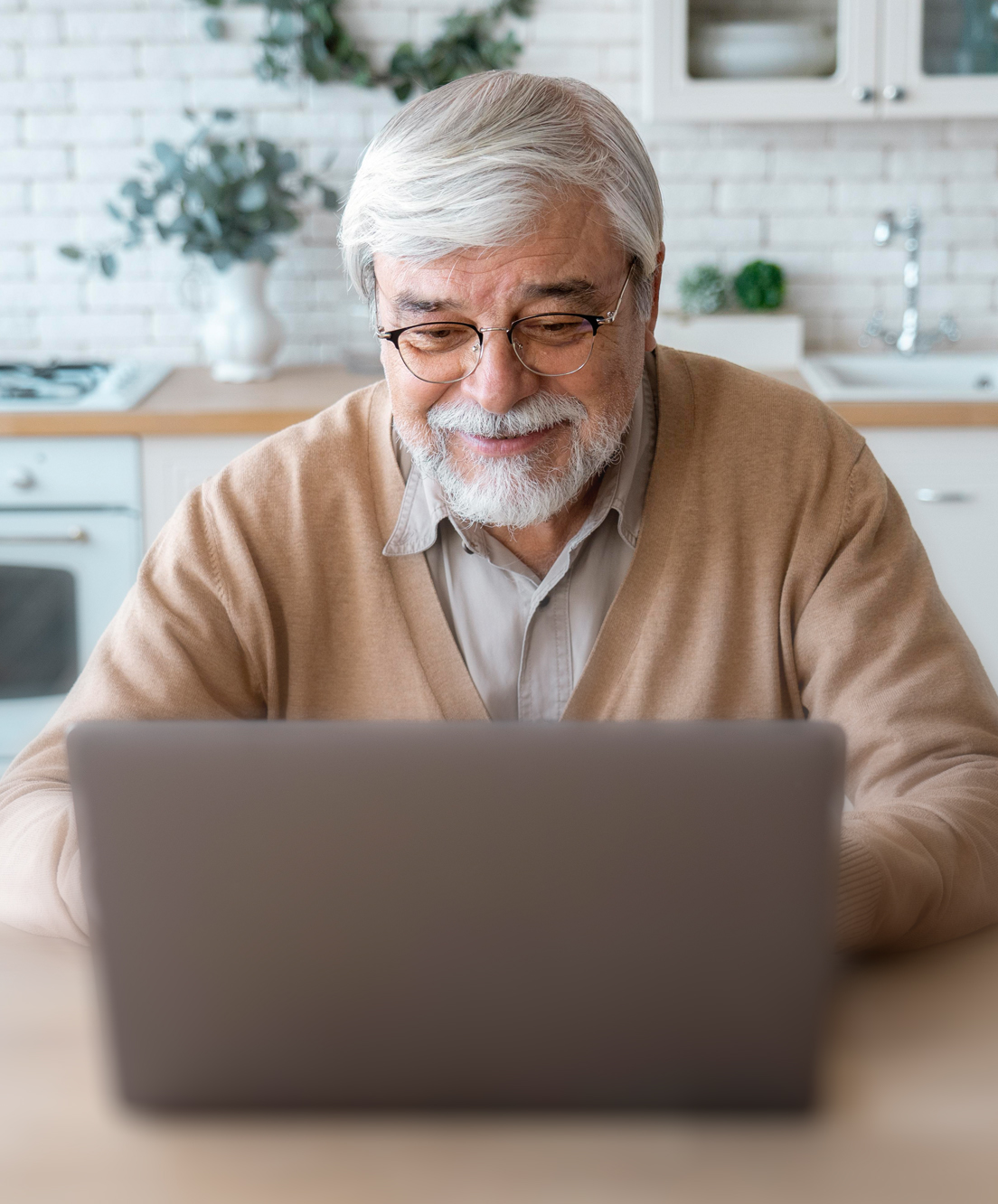 Smiling elderly man with glasses and white hair sitting at a table using a laptop in a kitchen.