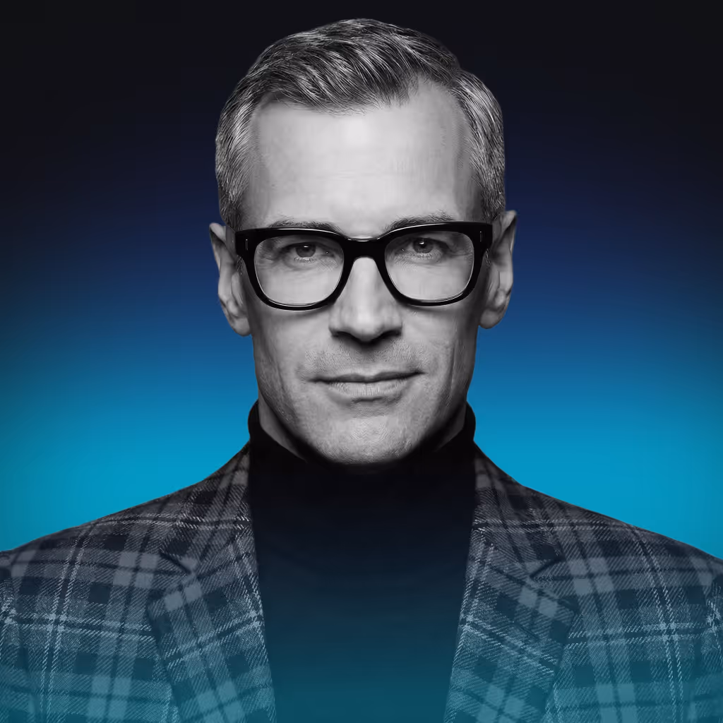 Portrait of a middle-aged man with short gray hair, black thick-rimmed glasses, wearing a checkered blazer and black turtleneck against a blue gradient background.