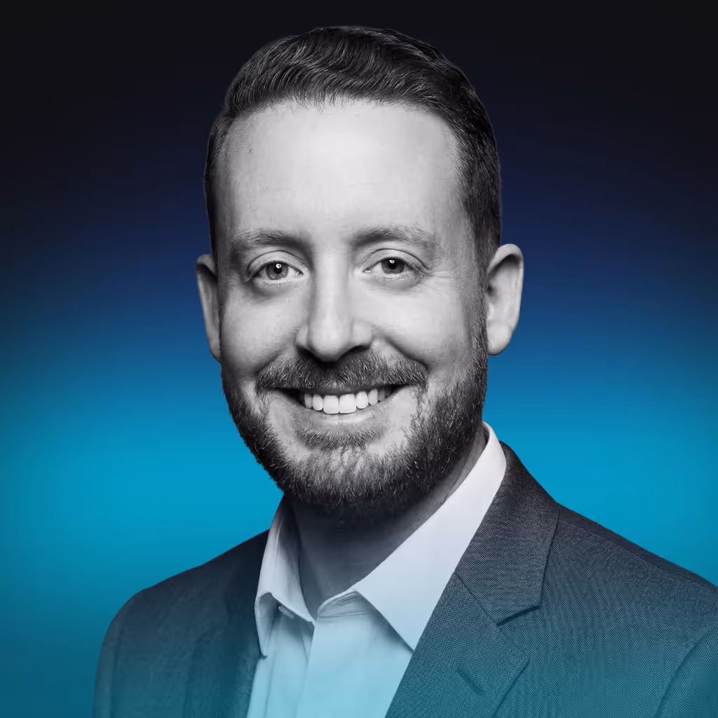 Smiling man with short hair and beard wearing a suit jacket and white shirt against a blue gradient background.