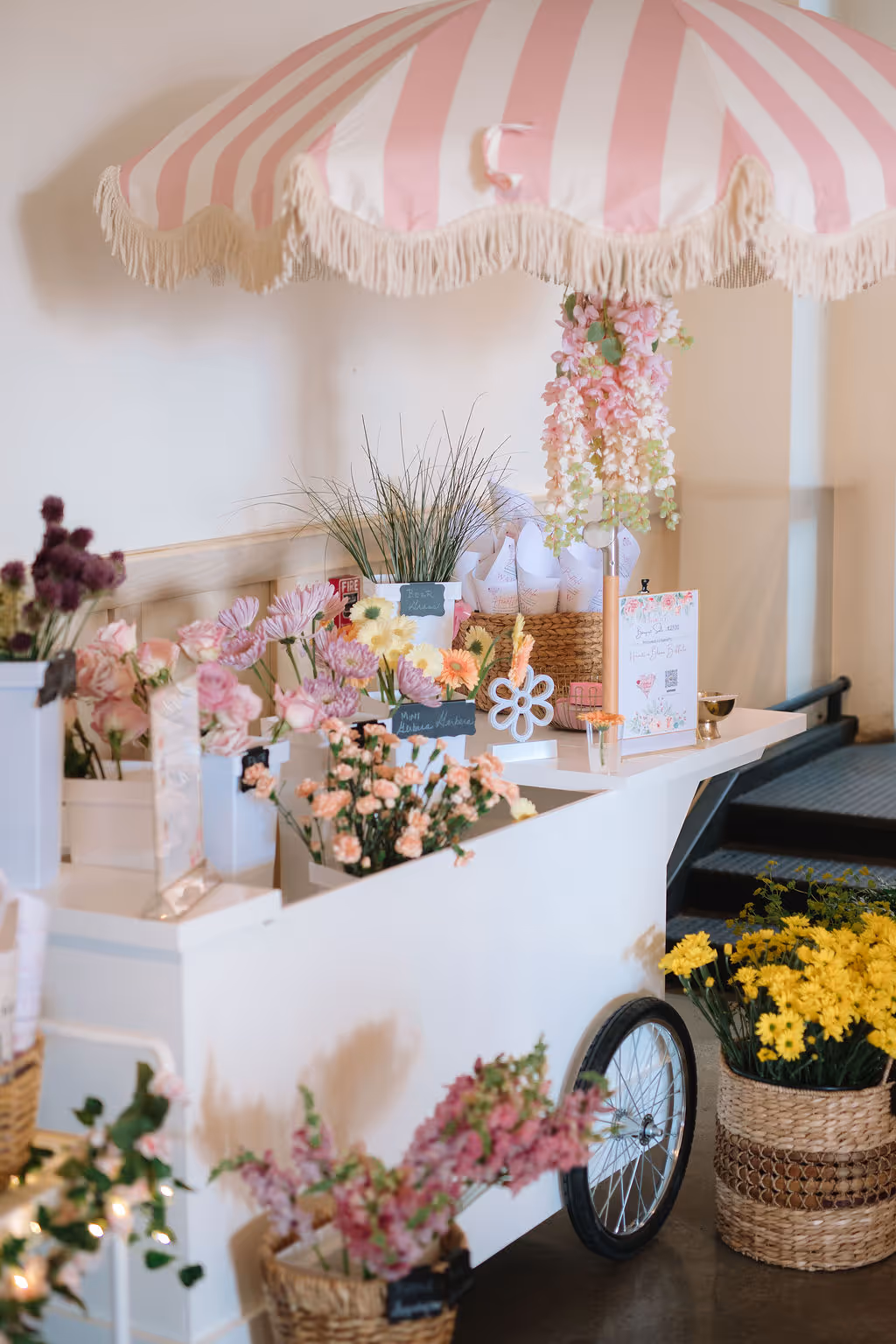 White flower cart with a pink and white striped canopy, displaying various bouquets and baskets of colorful flowers indoors.