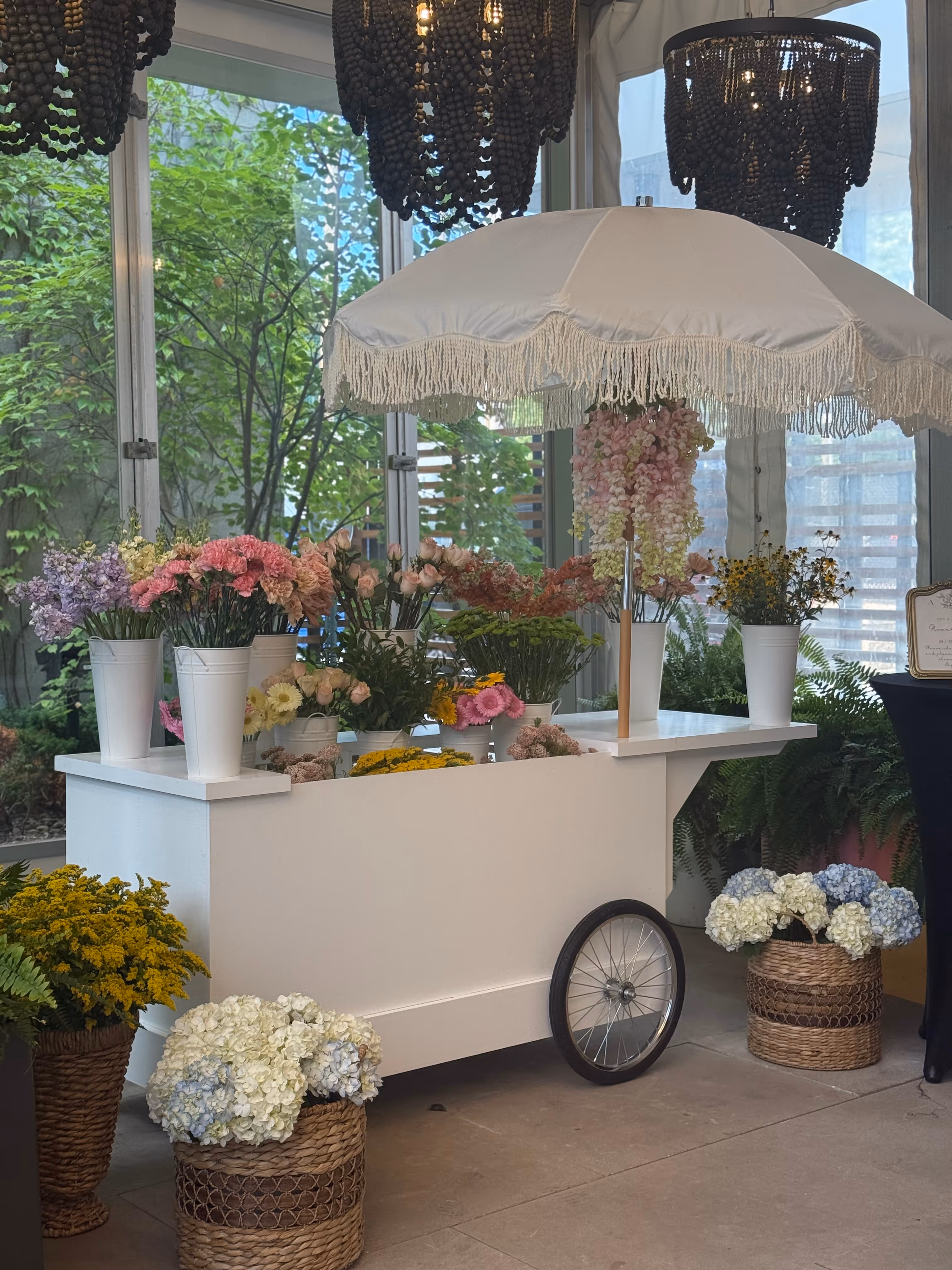 White flower cart with a fringed umbrella displaying various colorful flowers in white pots and wicker baskets indoors.