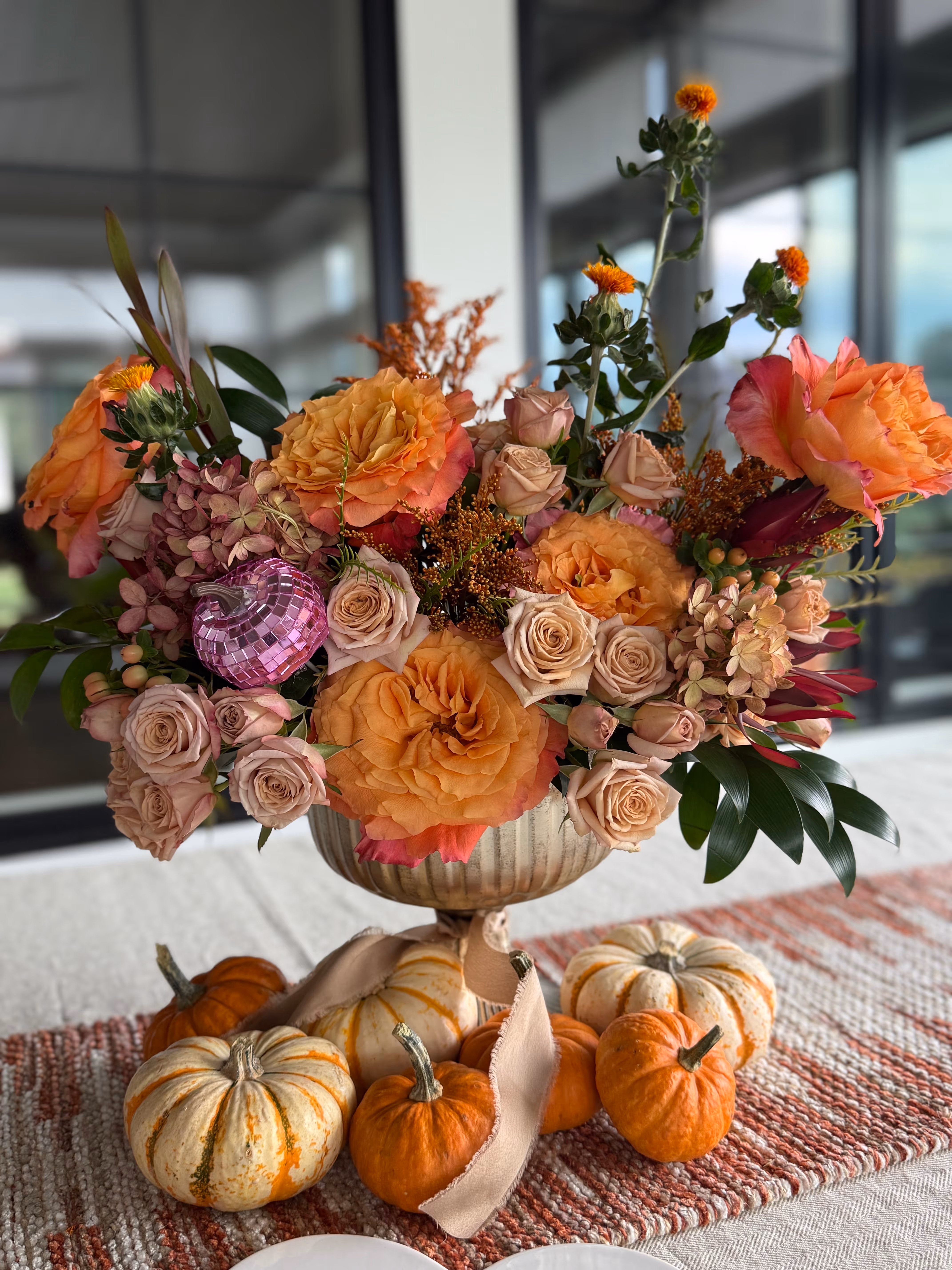 Autumn-themed floral arrangement with orange and beige roses, small orange flowers, green leaves, and a purple decorative ornament, surrounded by small orange and white pumpkins on a textured table runner.