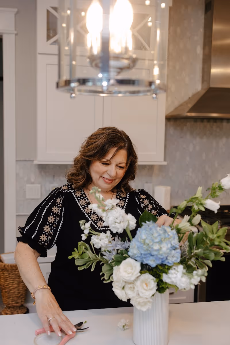 Woman arranging a bouquet of white and blue flowers in a white vase in a modern kitchen.