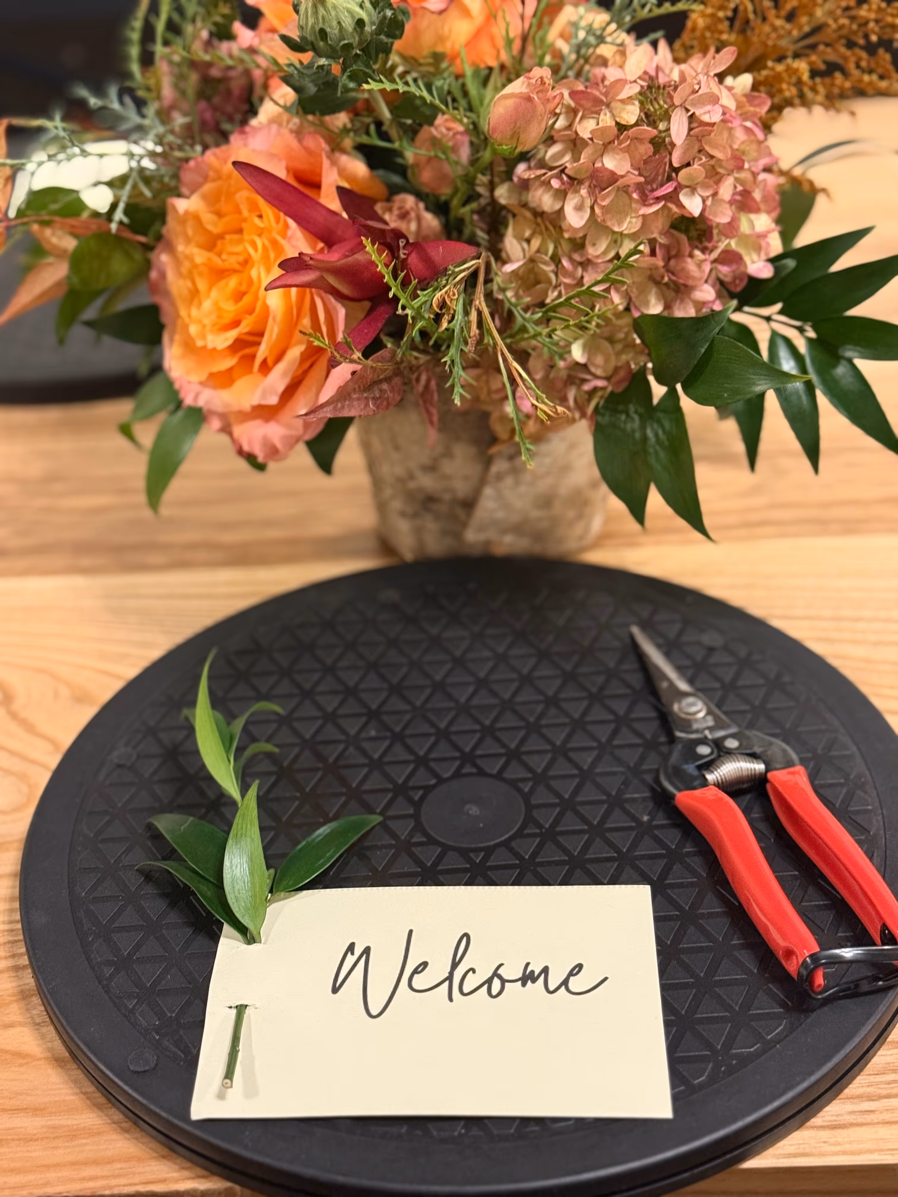 A floral arrangement with orange and pink flowers and greenery, a pair of red-handled pruning shears, and a cream card reading 'Welcome' on a black textured round mat on a wooden table.