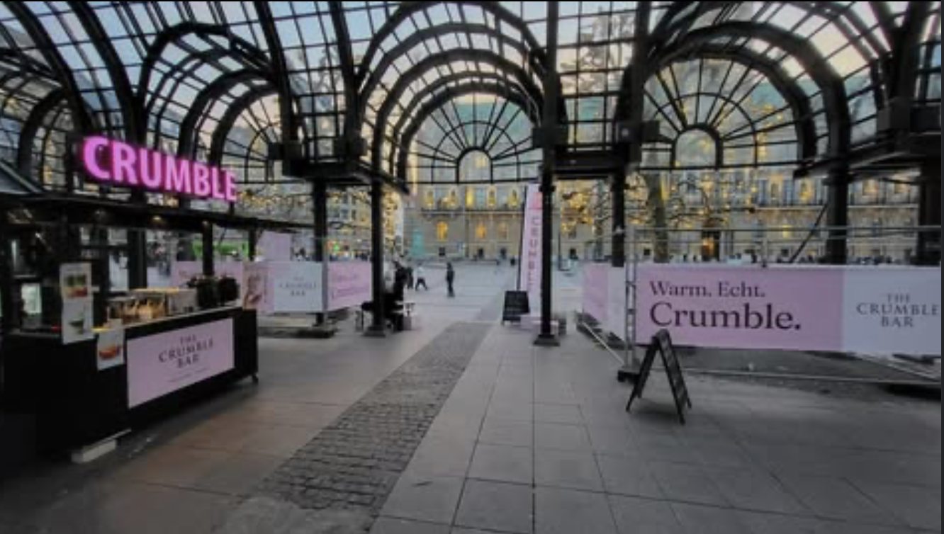 Outdoor covered area with a pink neon Crumble sign and pink banners advertising The Crumble Bar with a historic building in the background.
