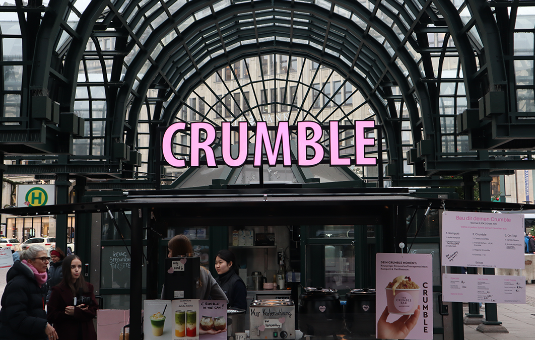 Street food stall under glass arch structure with pink illuminated sign reading 'CRUMBLE' and people gathered nearby.