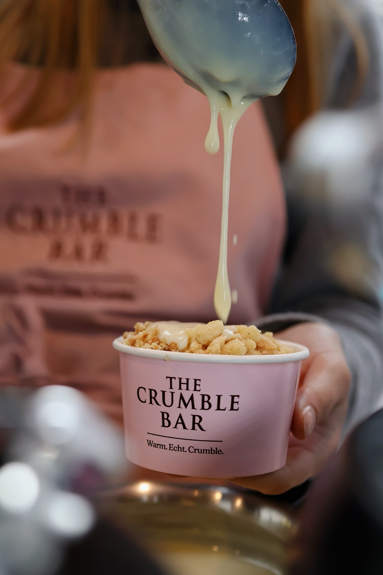 Hand holding a pink cup labeled 'The Crumble Bar' with crumbly dessert being drizzled with creamy sauce from a spoon.