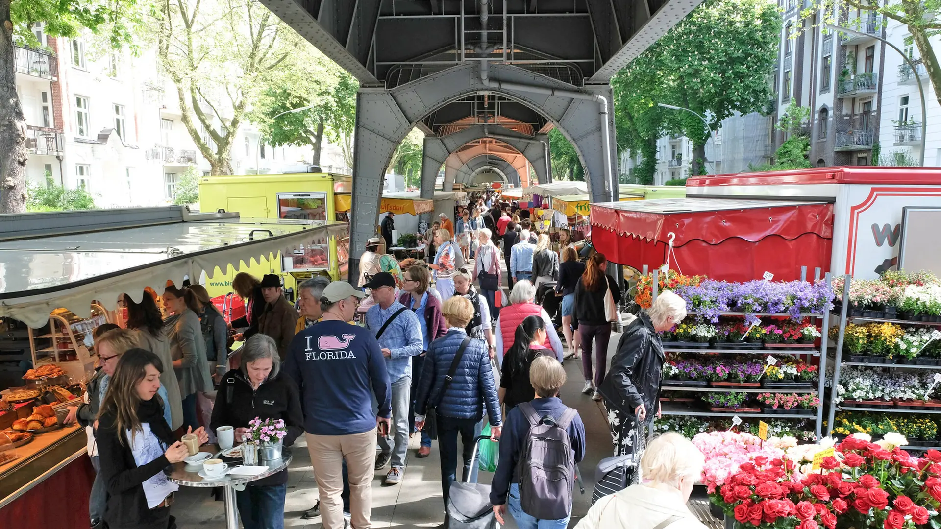 Crowded outdoor market under metal bridge with flower and food stalls and people walking and browsing.