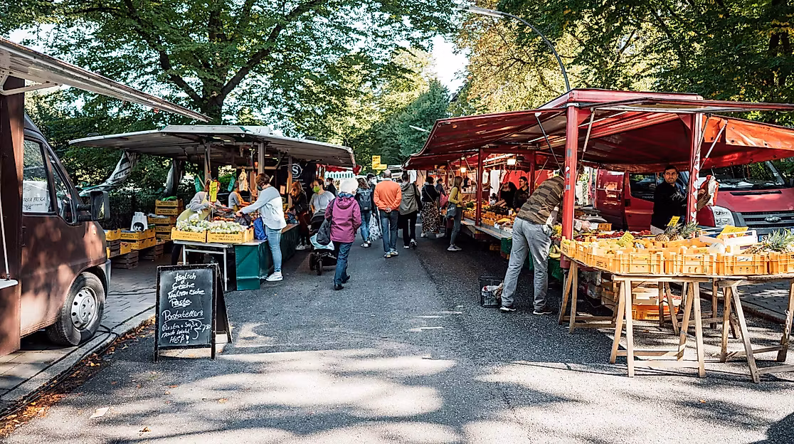 People shopping at an outdoor street market with stalls selling fruits and vegetables under trees.