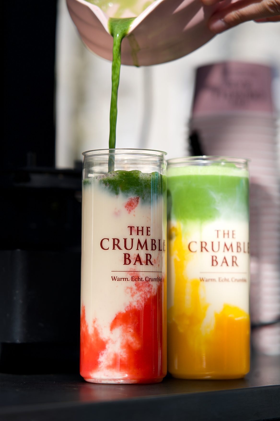 Two layered drinks with green liquid poured into one, in clear cups labeled The Crumble Bar.