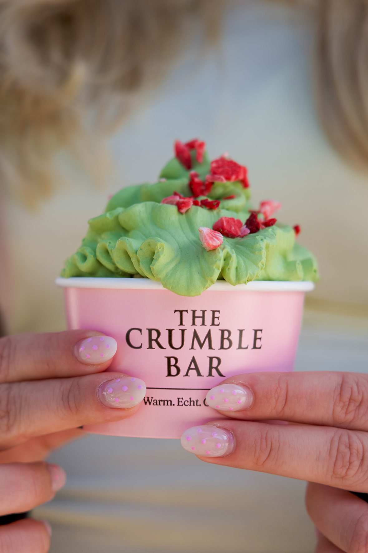 Hands hold a pink cup of green matcha ice cream with red sprinkles from The Crumble Bar.