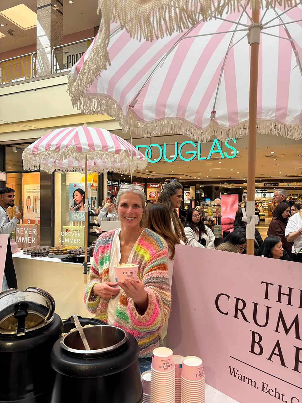 Smiling woman in a colorful sweater serving food in pink cups at The Crumble Bar under pink striped umbrellas.
