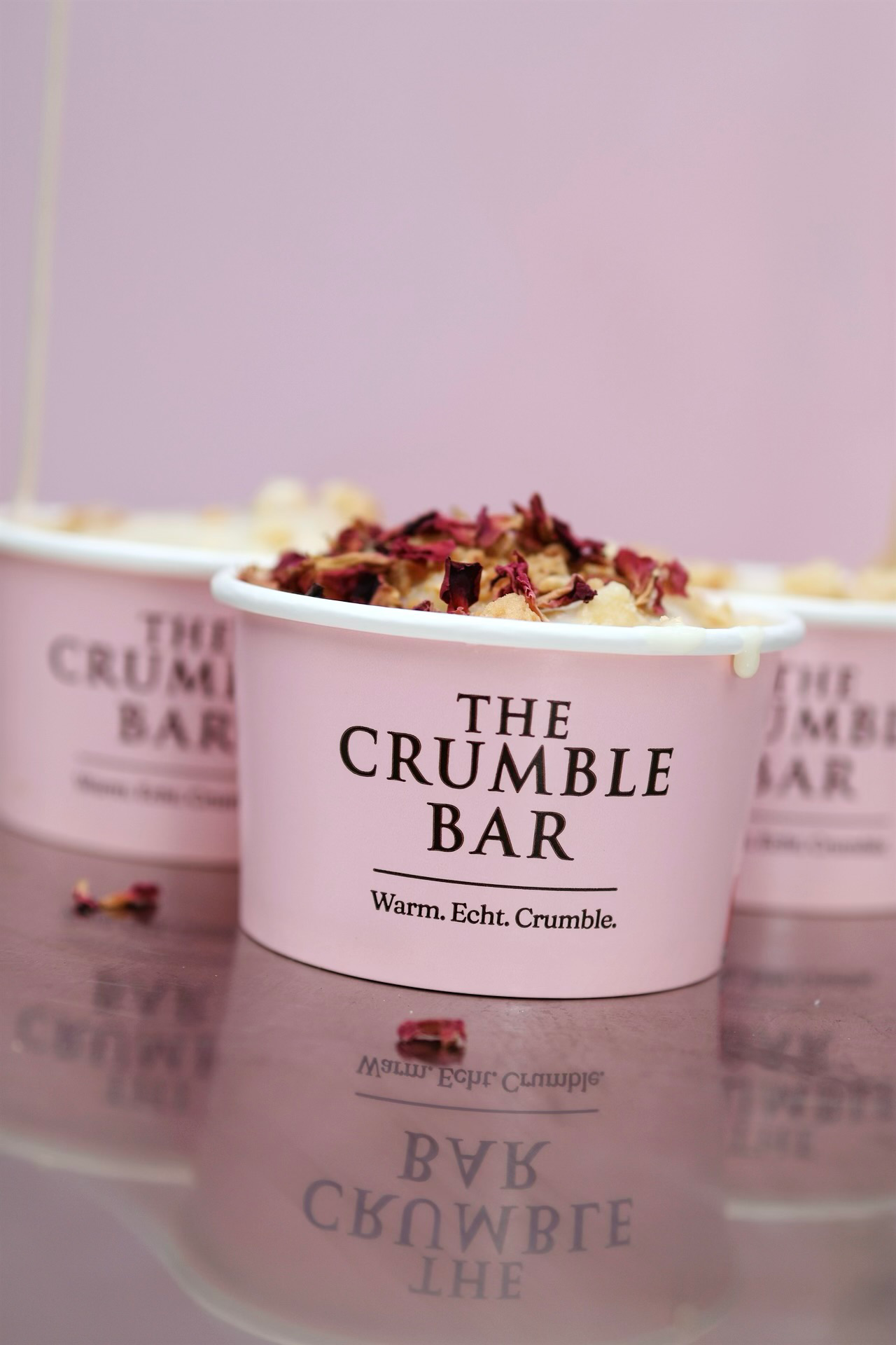 Pink cup labeled The Crumble Bar with crumble dessert topped with red flower petals.