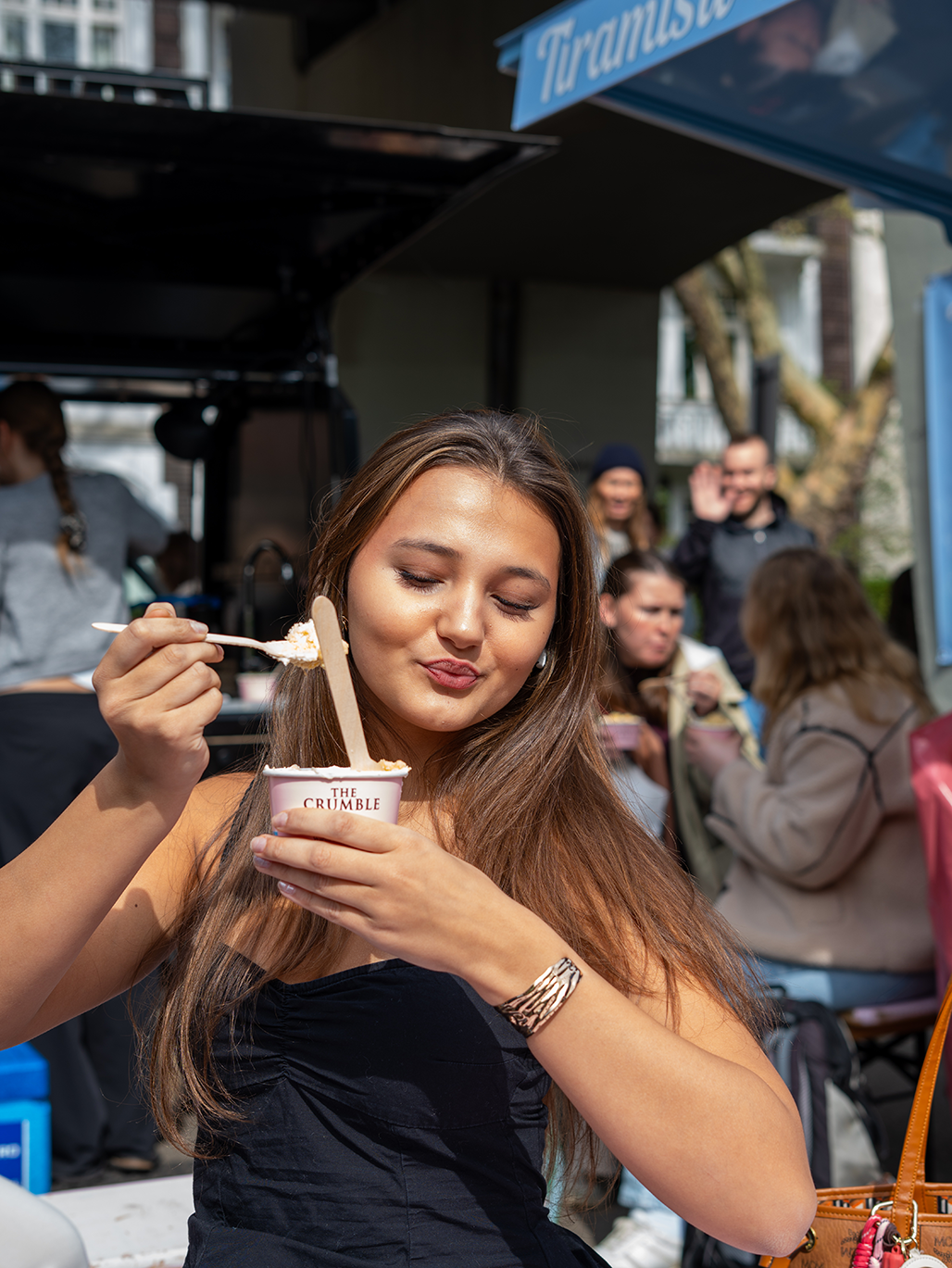 Woman enjoys a cup of dessert from The Crumble, holding a spoon and cup with a satisfied expression.
