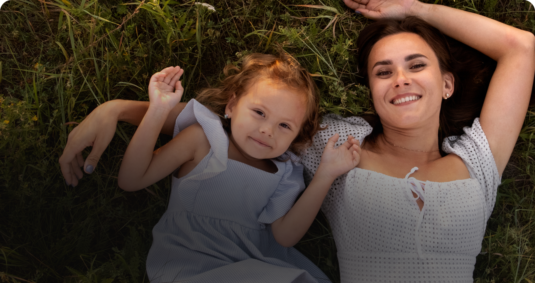 Smiling woman and young girl lying on grass outdoors, both wearing light dresses.