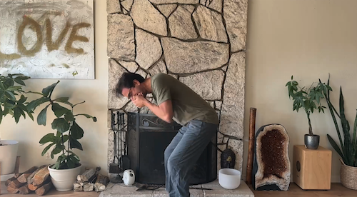 Man in casual clothes leaning forward and covering his face with his hands in a cozy room with stone fireplace, potted plants, and a decorative amethyst geode.