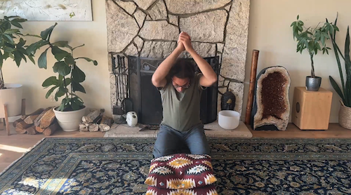 Person kneeling on patterned rug, about to pound a colorful pillow with clasped hands in a cozy room with plants and a stone fireplace.