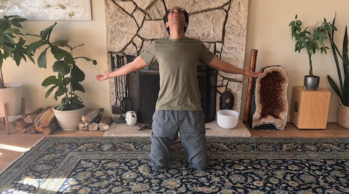 Man kneeling on a patterned rug with arms outstretched and head tilted back in front of a stone fireplace.