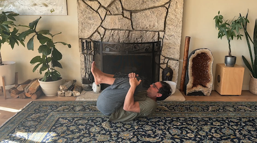 Man lying on a carpet in front of a stone fireplace, hugging his knees in a yoga pose, surrounded by plants and decorative items.