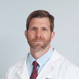 Professional middle-aged male doctor with short brown hair and beard wearing a white lab coat, light blue shirt, and maroon tie against a plain light gray background.