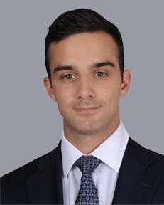 Portrait of a young man with short dark hair wearing a white shirt, patterned tie, and dark suit jacket against a gray background.