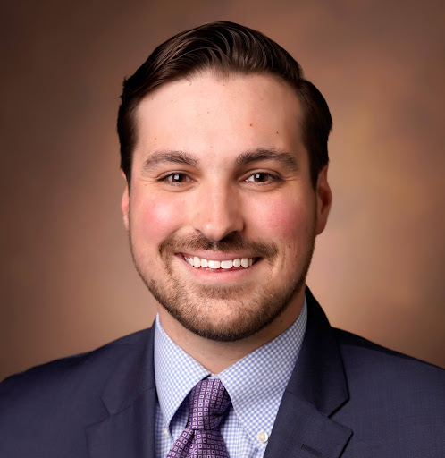 Smiling man with neatly combed dark hair, wearing a navy suit, checked shirt, and patterned tie against a brown gradient background.