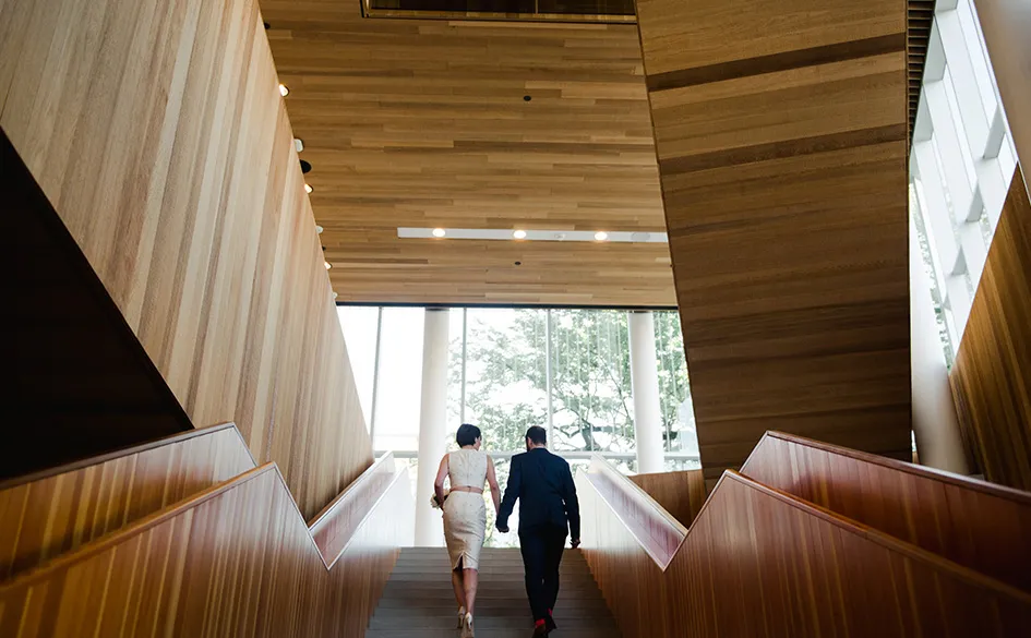 Couple holding hands walking up wide wooden stairs inside modern building with large windows.