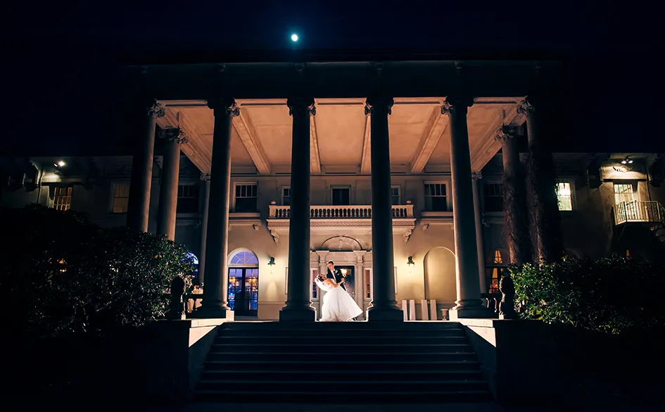 Bride and groom dancing in front of a grand building with tall columns at night under a full moon.