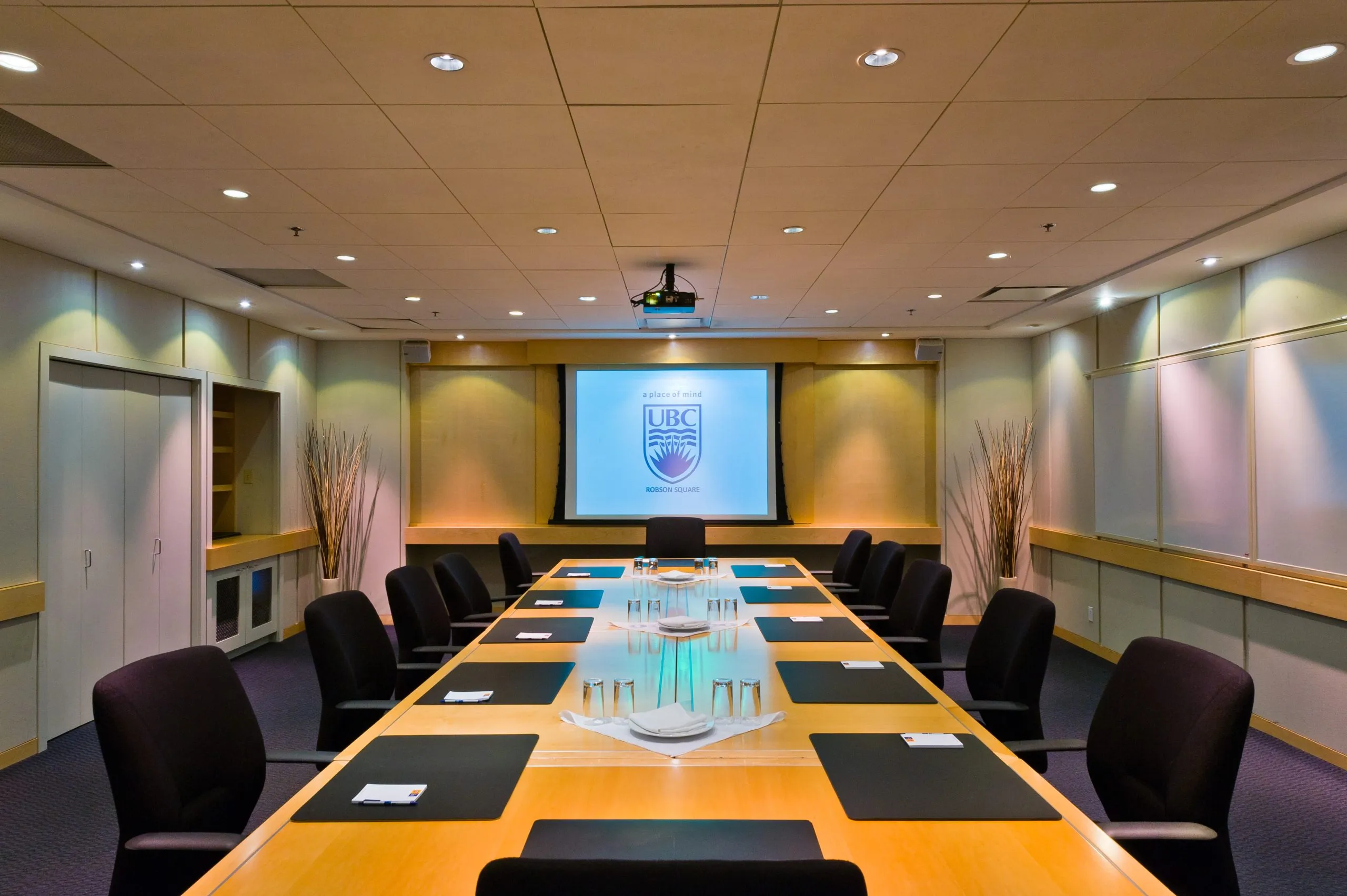 Modern boardroom with long wooden table, black chairs, place mats, glassware, and a UBC projection screen at the front.