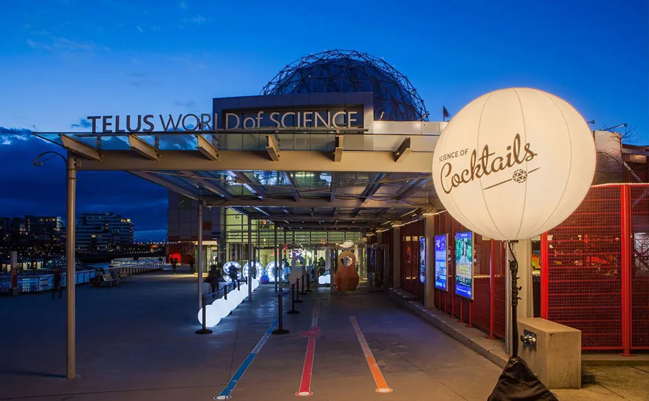 Entrance to TELUS World of Science at dusk with illuminated 'Science of Cocktails' balloon and cityscape in the background.