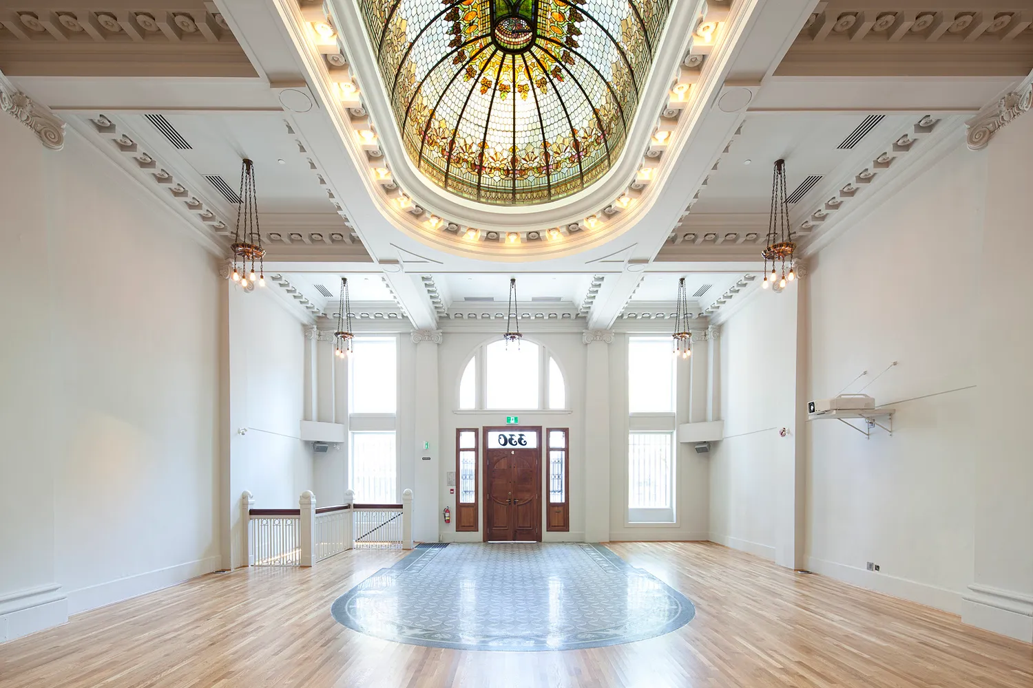 Spacious room with polished wooden floors, stained glass ceiling dome, white walls, and wooden double doors flanked by tall windows.