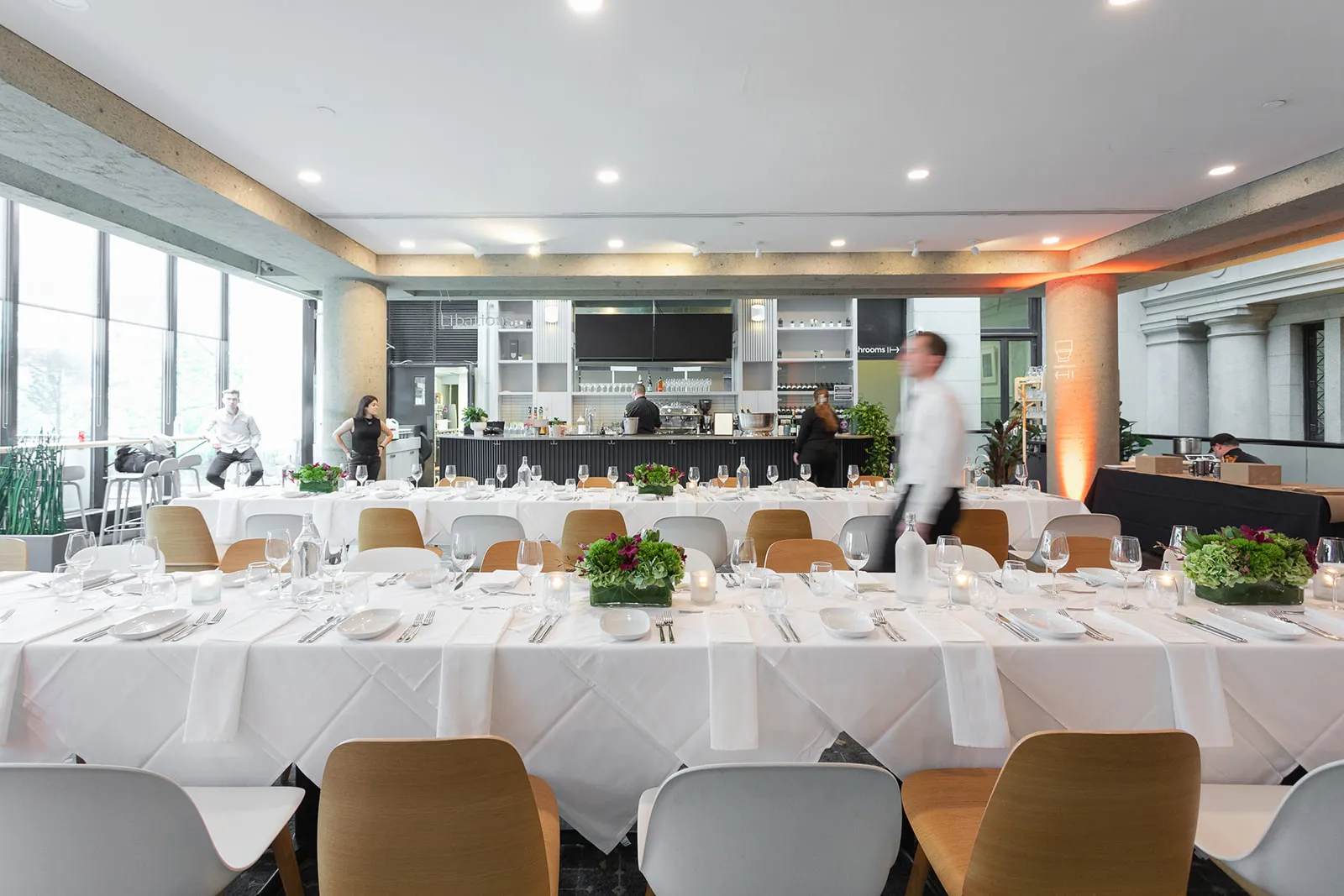 Bright modern dining area with long white-clothed tables set for a formal meal, decorated with green and purple floral centerpieces, and staff preparing in the background.