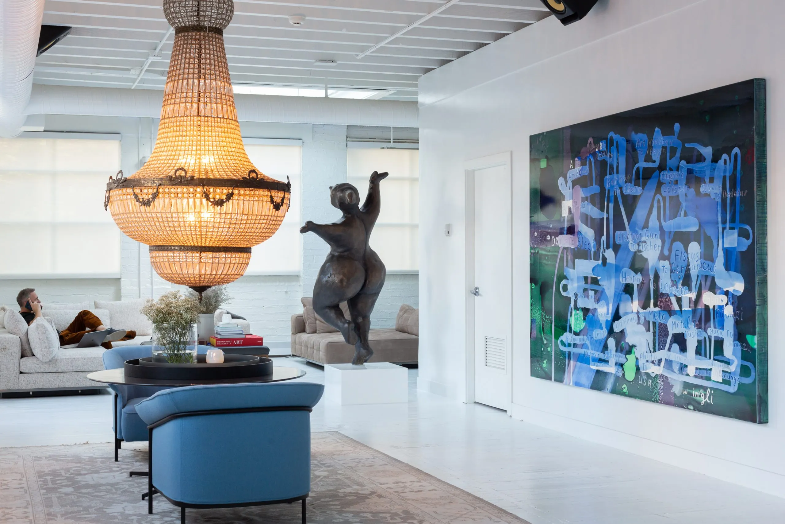 Modern living room with large chandelier, abstract blue painting on wall, bronze sculpture, blue chairs, and beige sofas.