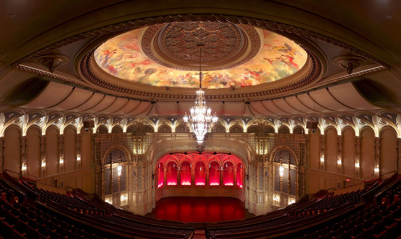 Ornate theater interior with a large sparkling chandelier, colorful painted dome ceiling, and red-lit stage.