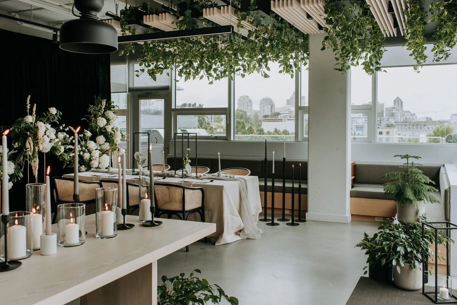 Elegant dining area with white candles on tables, green plants hanging from ceiling, white floral arrangements, and large windows overlooking a cityscape.