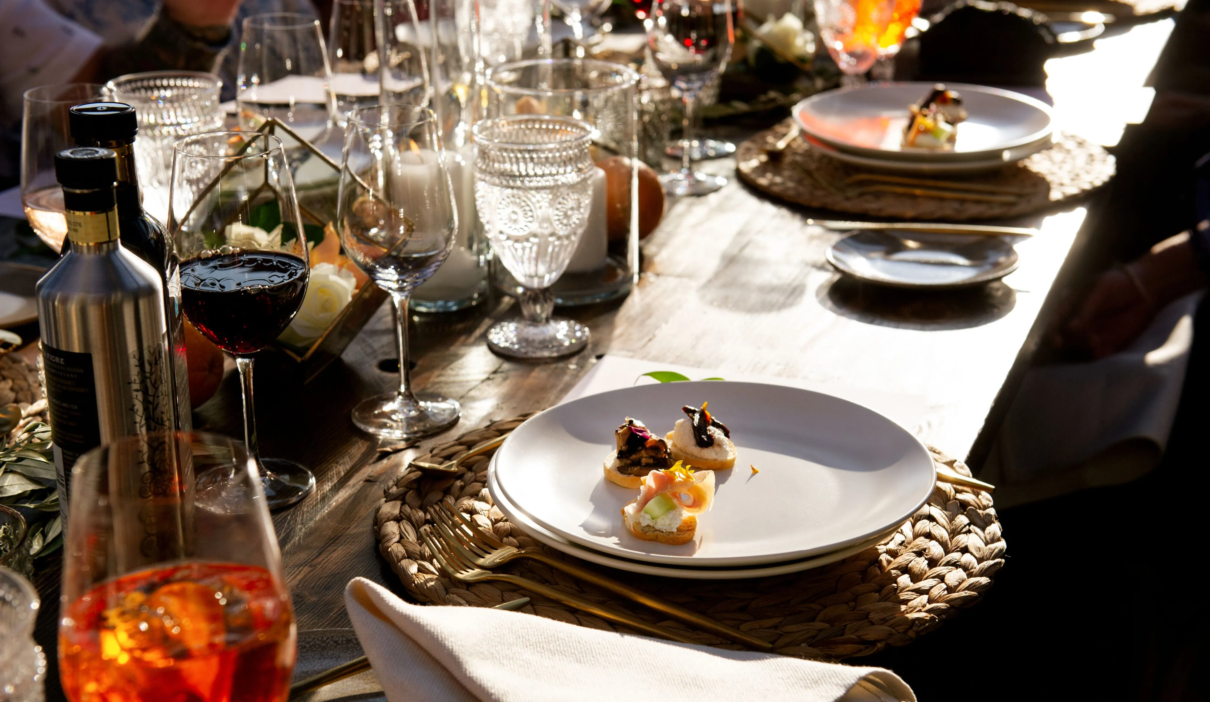 Elegant table setting with white plates holding gourmet bite-sized appetizers, surrounded by wine glasses, candles, and decorative elements on a wooden table.