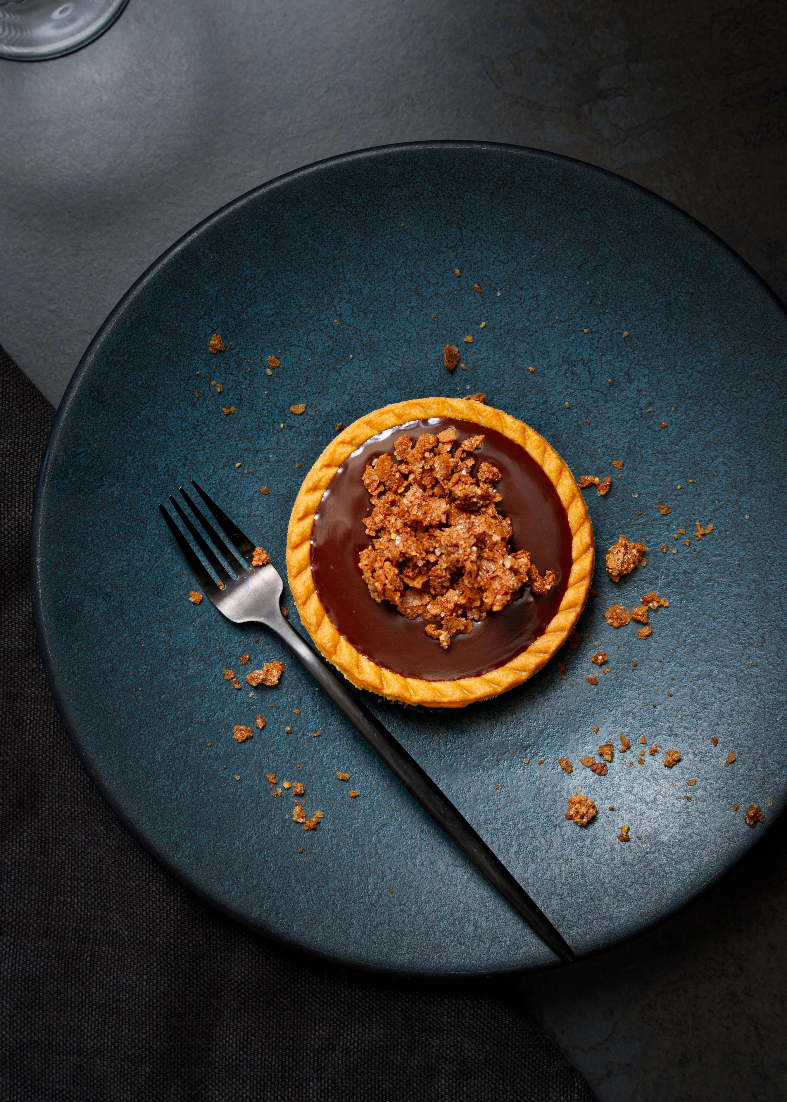 Chocolate tart with crumb topping on a dark blue plate with a black fork and scattered crumbs.