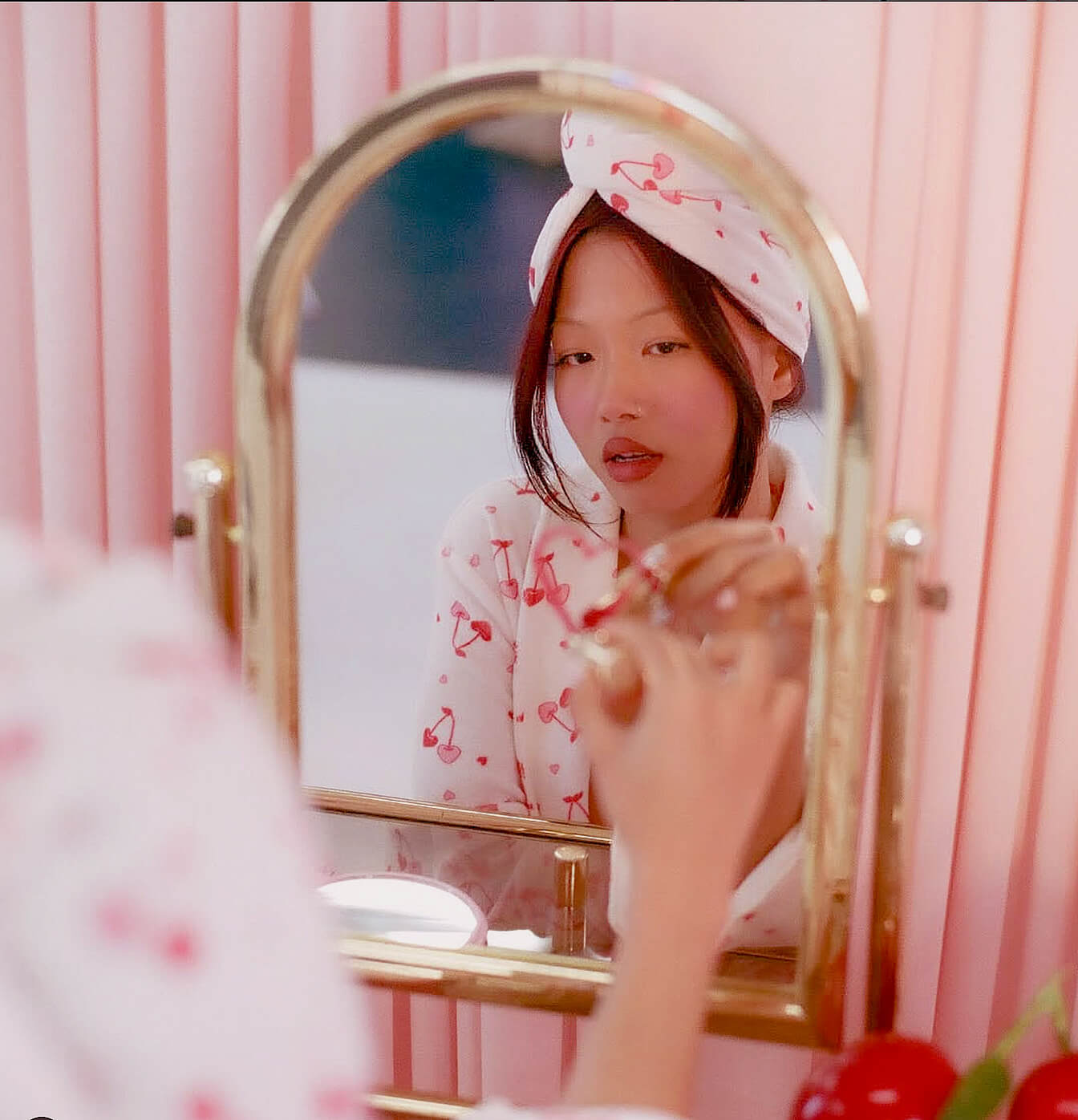 Woman wearing a cherry-patterned headband and robe looking into a gold-framed mirror with a pink background.