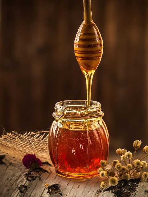 Honey dripping from a wooden honey dipper into a glass jar of honey on a rustic wooden surface with dried flowers nearby.