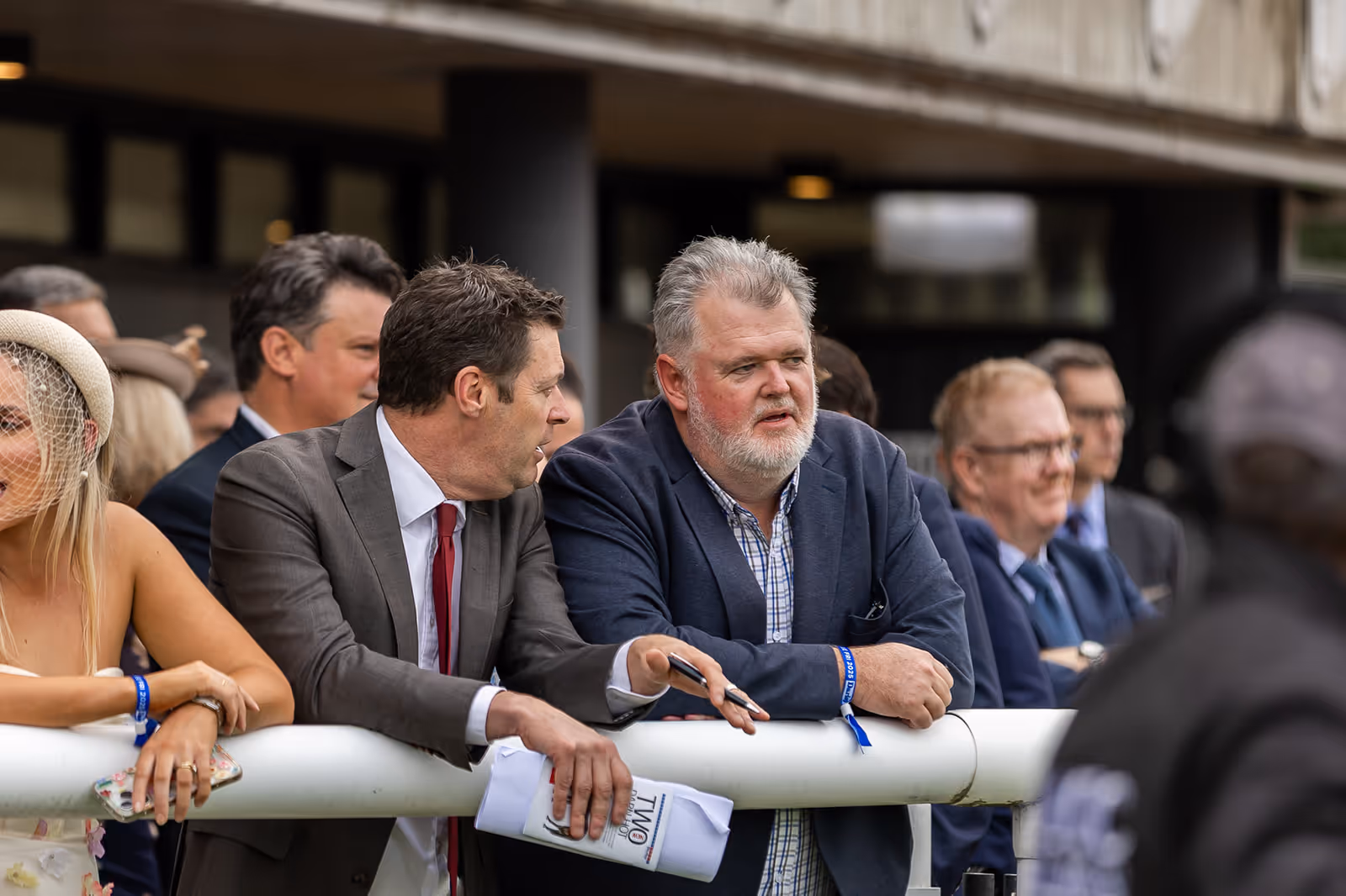 Two men in suits leaning on a white railing, engaged in conversation, with other people in the background.