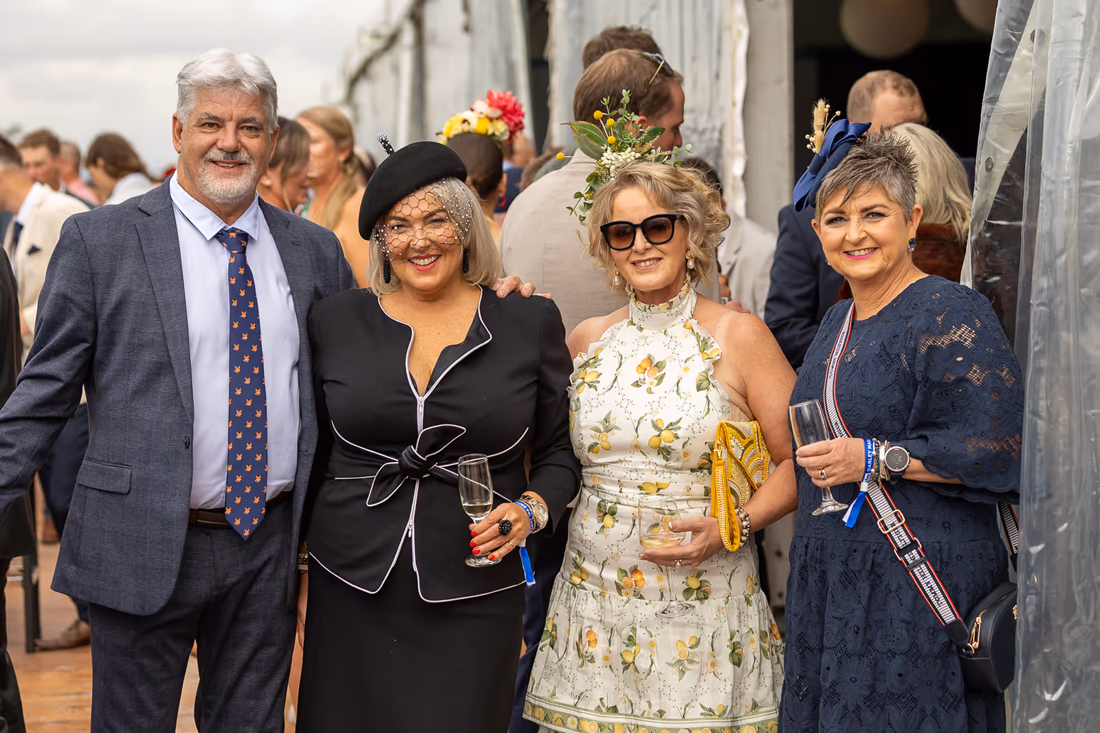 Four well-dressed adults smiling at a social event, two women wearing decorative hats and holding drinks.