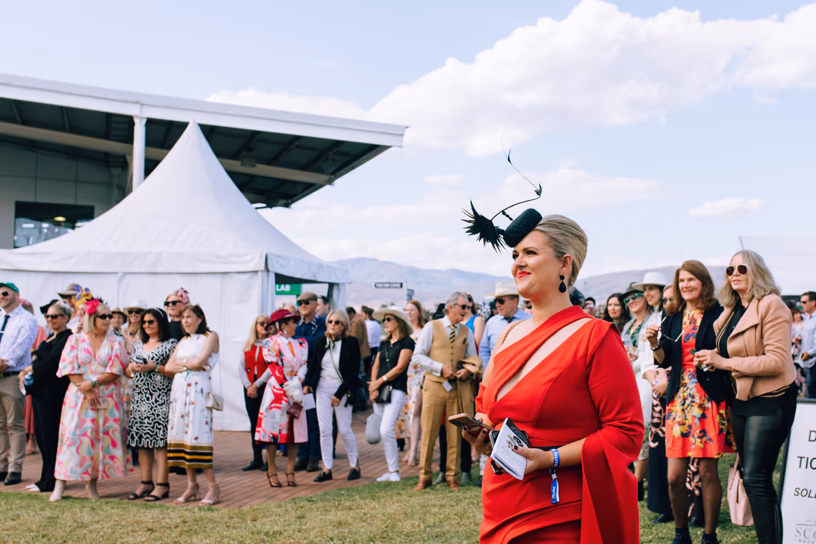 Woman in a stylish red dress and black feathered fascinator at an outdoor event with a crowd and white tents in the background.