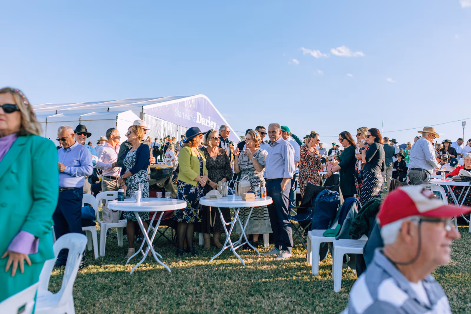 People socializing outdoors at an event with round white tables and a large white tent in the background under clear blue sky.