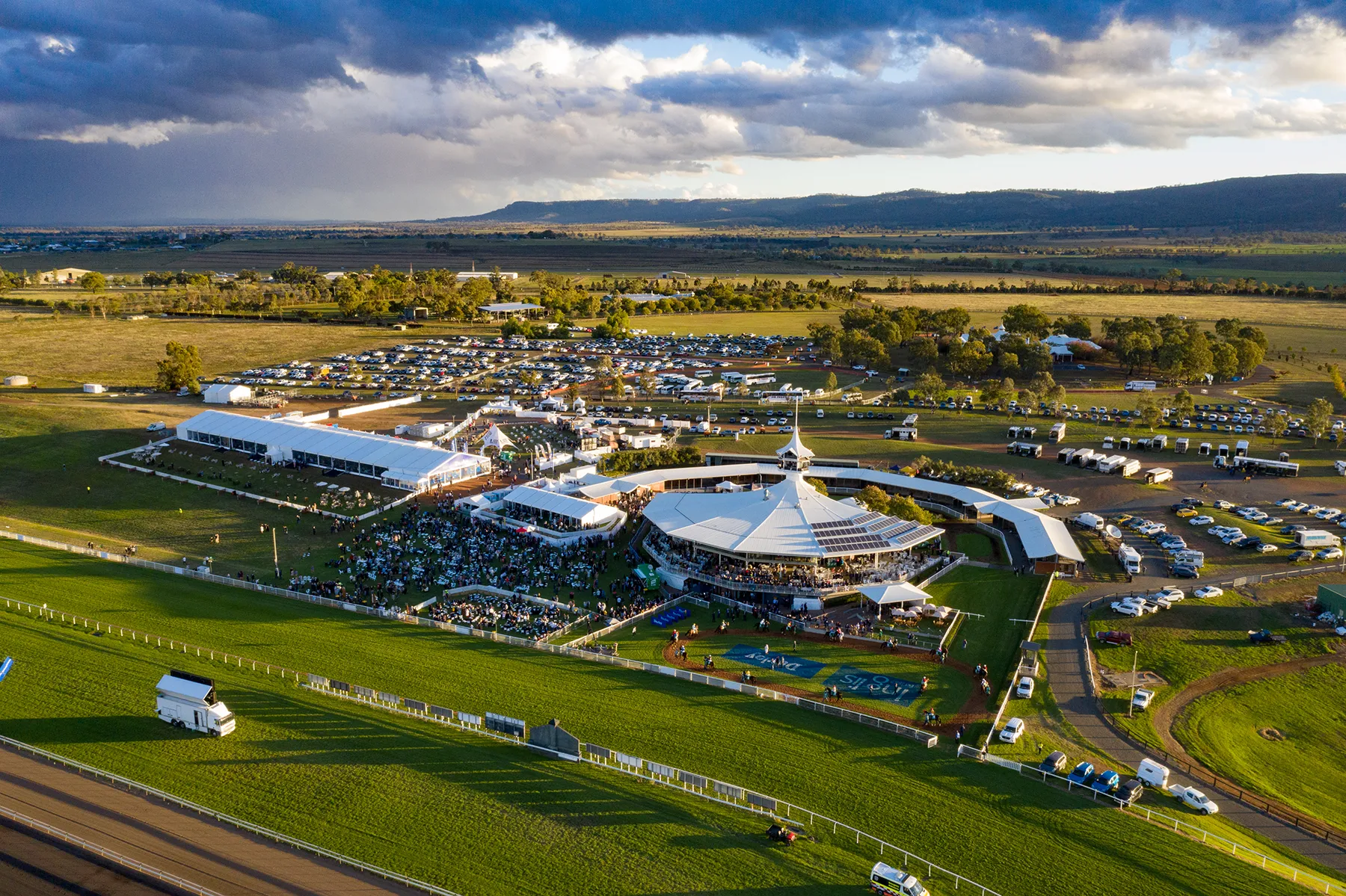 Aerial view of a racecourse event with a crowd gathered around white tents and a central pavilion, surrounded by parked cars and green fields under a partly cloudy sky.