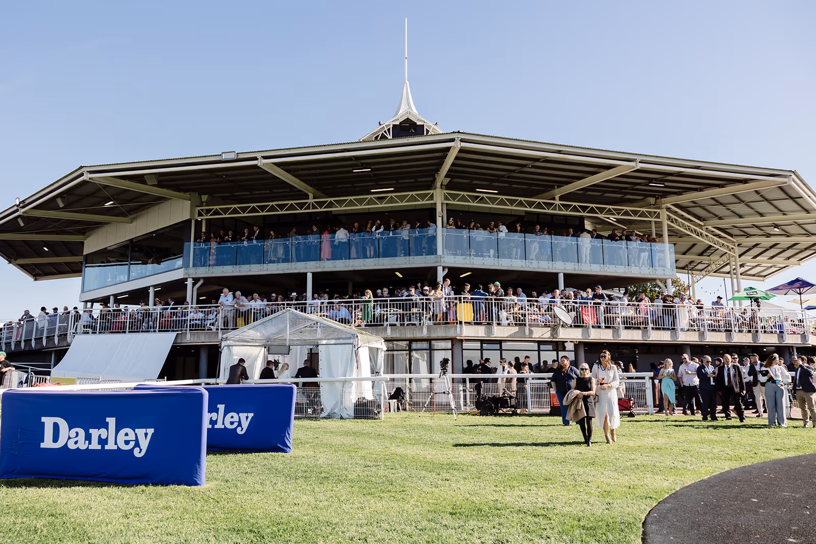 Crowd of people standing and walking near and on a two-story grandstand at a daytime outdoor event on green grass.