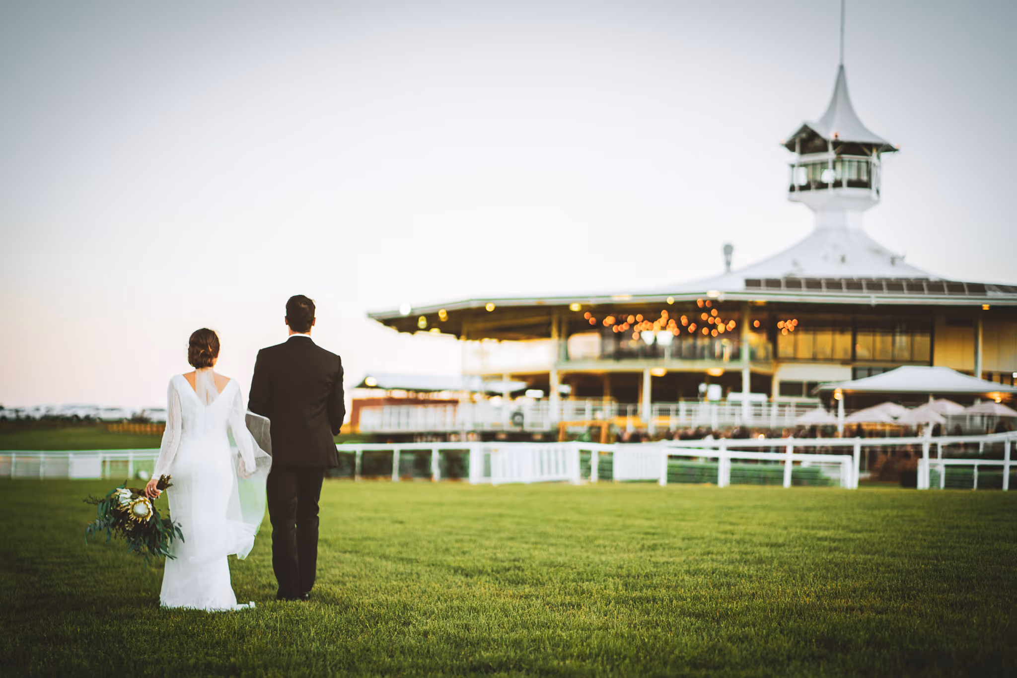 Bride and groom walking hand in hand across a green lawn toward a lit building with a white tower at dusk.
