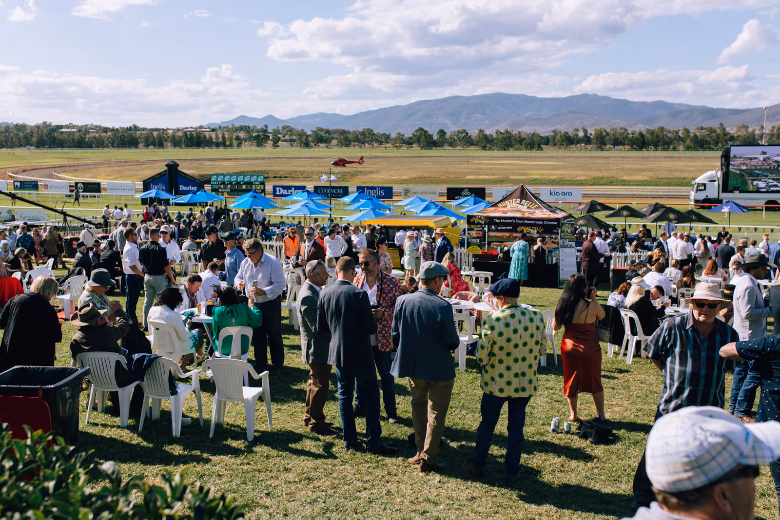 Crowd of people socializing with drinks and food under blue and black umbrellas at a sunny outdoor event near a racetrack with mountains in the background.